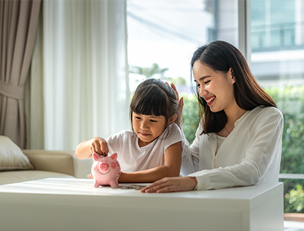 Happy mother and daughter saving money putting coins into piggy bank on table at home.