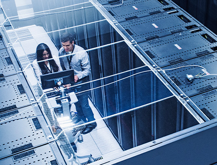 Two tech industry professionals stand in between a row of server racks while analyzing data on a computer.