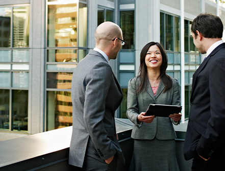 Three professionals are networking outside of a business building while the female professional smiles at her two male colleagues holding a digital tablet.