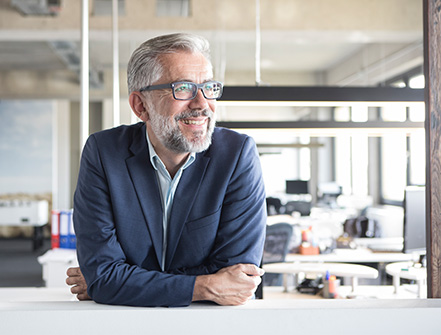 A middle-aged man with glasses and grey hair smiles as he leans over a window inside his small business office. 