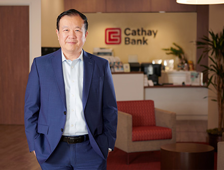 Chang M. Liu se para dentro de una sucursal de Cathay Bank con un traje azul oscuro y sonríe a la cámara.