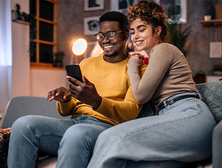 Una pareja joven se sienta en un sofá de la sala de estar y sonríe a su teléfono móvil mientras revisa sus opciones de hipoteca de Cathay Bank.