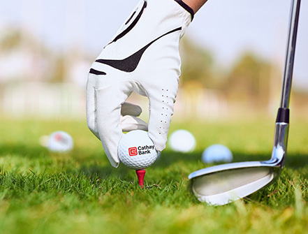 A golf ball with Cathay Bank logo sits outdoors on grass with a golf club and hand next to it for the annual tournament in California. 