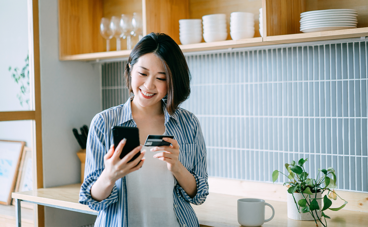 A woman is standing in a modern kitchen holding a smartphone in one hand and a credit card in the other, suggesting an online payment or mobile banking activity.