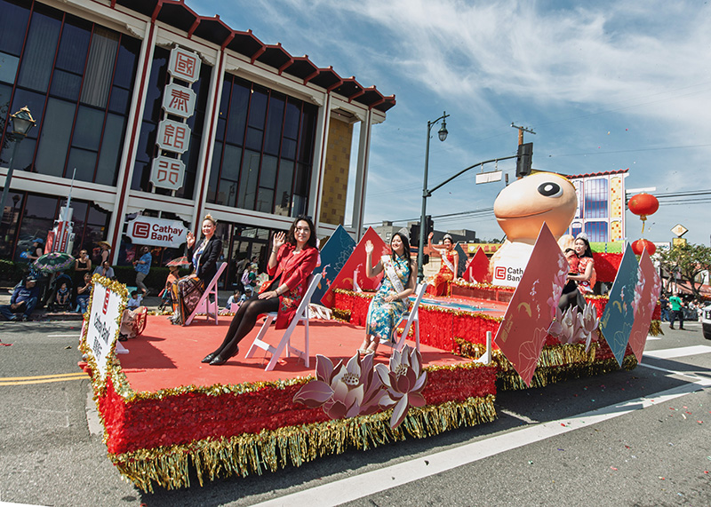 A parade float shows a Cathay Bank logo for the Year of the Snake celbration with a large snake statue as various women from the local community wave to the crowd.