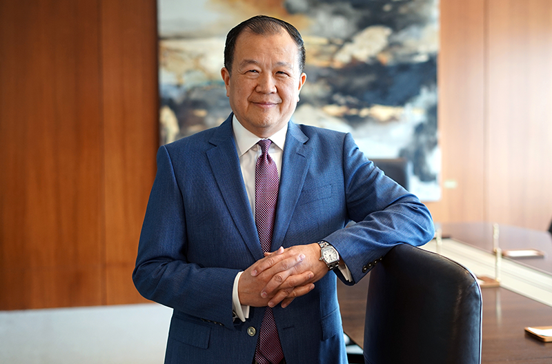 Chang M. Liu, presidente y director ejecutivo de Cathay Bank, vestido con un traje azul de negocios, camisa blanca y corbata, aparece de pie en una oficina moderna; tiene las manos entrelazadas y apoyadas en una silla, con una pared revestida de madera y un cuadro abstracto al fondo.
