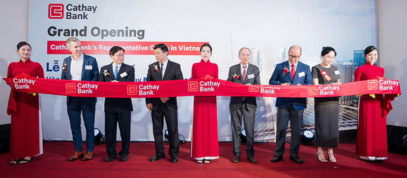 Ceremonia de inauguraci&oacute;n de la oficina de representaci&oacute;n de Cathay Bank en Vietnam, con los asistentes sosteniendo una cinta roja con el logotipo de Cathay Bank frente a un tel&oacute;n de fondo de la entidad.