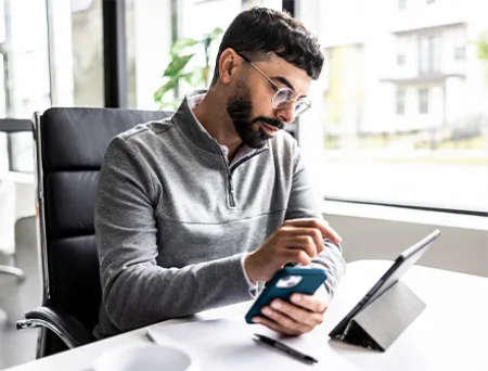 A man in a grey quarter-zip sweater and glasses sits at his work desk. In his left hand he is holding a phone, while his right hand is focused and pointing toward an iPad lying on the desk in front of him. 