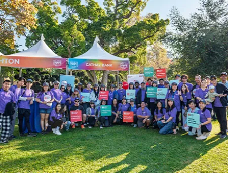 Cathay Bank team members in purple t-shirts stand together in an outdoor grass area with a Cathay Bank tent behind them, holding Cathay Bank signs for a group photo.