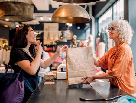 Client and retail worker exchange a shopping bag across a boutique clothing store counter, with racks of apparel and warm lighting in the background.