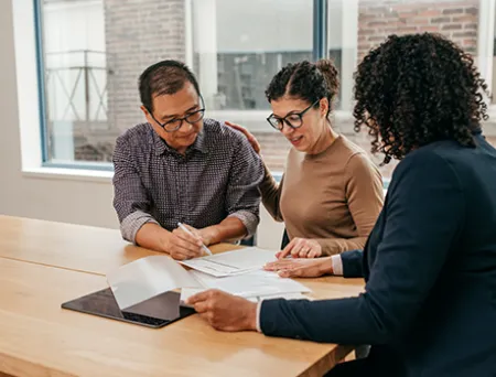 Three adults sit at a table in a bright office, reviewing and signing documents together, with a tablet nearby and large windows in the background.