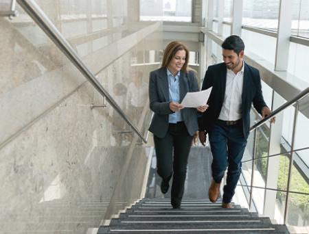 A businesswoman and her male colleague walk up the stairs at an open space office while looking at a business LOC document.