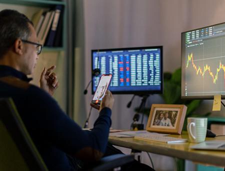 Un hombre de negocios sostiene su teléfono celular para chatear por video a un cliente en el extranjero mientras se sienta frente a su escritorio, viendo las estadísticas de divisas en la pantalla del ordenador.