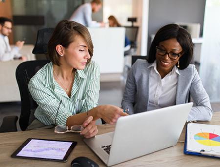 Two business women chat over international trade finance while looking at a laptop in an office.