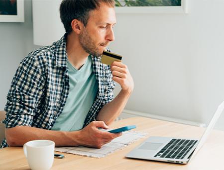 A man sits at his wooden desk in a brightly lit office while staring at his laptop and holding his credit card with a worried look about fraud alerts