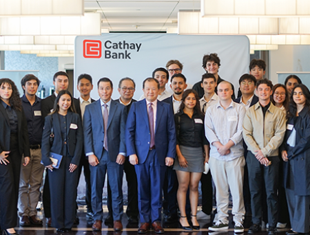 Un numeroso grupo de estudiantes y representantes de Cathay Bank posan juntos frente a un fondo con el logotipo de Cathay Bank. El grupo viste ropa formal o semiformal de negocios y posa para una foto grupal formal dentro de las oficinas corporativas, con iluminación y decoración interior modernas de fondo.