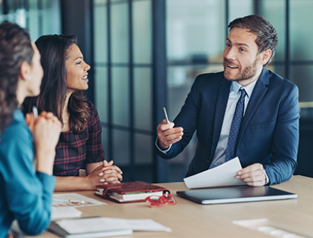 Three professionals in a modern office discuss advanced banking strategies, with documents, a notebook, and a laptop on the table during a collaborative meeting.