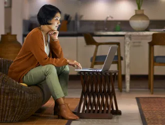 A woman with short black hair sits on a couch at home while trying to open an online banking account using her laptop.