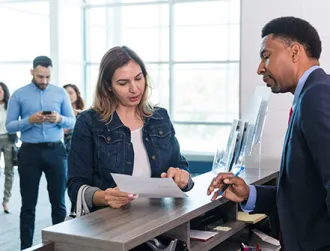 An adult male bank manager stands at the counter to answer the adult woman's questions about her monthly statements and recent check cashing.