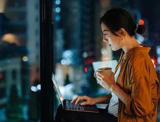 A woman in an orange shirt sits in front of her laptop in a dimly lit apartment, checking to report a phishing scam online.