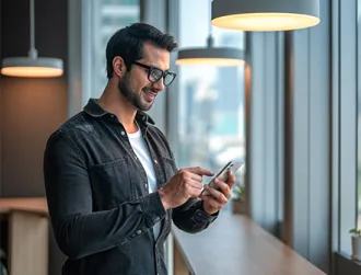 A male wearing glasses in a well-lit office space with warm lamps hanging from the ceiling stares happily at his phone while receiving security protection alerts from his mobile banking app.