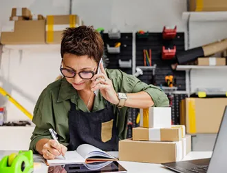 An employee at a small business wearing a green blouse with black overalls smiles as she speaks with a customer on the phone over her office desk.