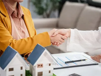 Two individuals sit at a table and shake hands over a mortgage refinancing agreement, with miniature house models, a clipboard of loan documents, and a pen on the table, symbolizing a completed home refinance deal.