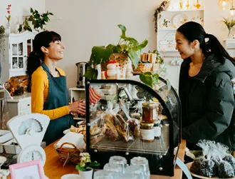 Small business owner and their shop customer converse at a café counter with a glass pastry display, surrounded by plants and warm lighting.