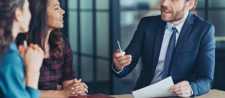 Three professionals in a modern office discuss advanced banking strategies, with documents, a notebook, and a laptop on the table during a collaborative meeting.