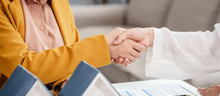 Two individuals sit at a table and shake hands over a mortgage refinancing agreement, with miniature house models, a clipboard of loan documents, and a pen on the table, symbolizing a completed home refinance deal.