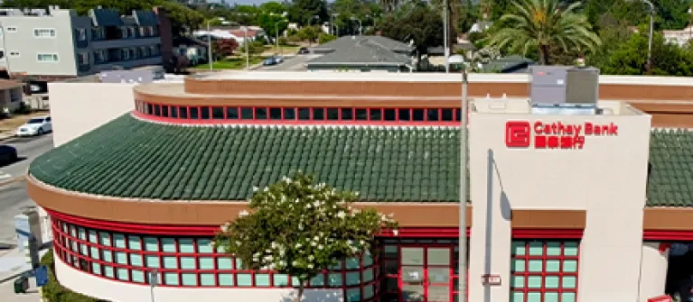 Exterior view of Cathay Bank’s Alhambra Branch, featuring a cream-colored building with a green tile roof, red-framed windows, and prominent Cathay Bank signage. 