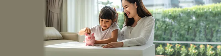 Happy mother and daughter saving money putting coins into piggy bank on table at home.
