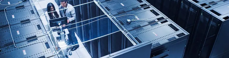 Two tech industry professionals stand in between a row of server racks while analyzing data on a computer.