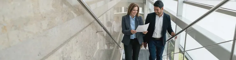 A businesswoman and her male colleague walk up the stairs at an open space office while looking at a business LOC document.