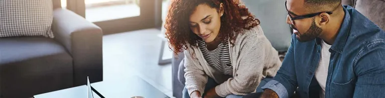 A young couple drinks coffee and goes over their finances while sitting in the living room of a cozy home.