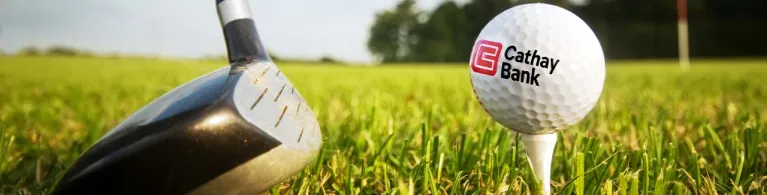 A Cathay Bank Golf Ball sits on the grass at an outdoor golf course with a golf club behind it.