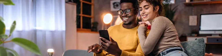 A young couple sits on a living room couch and smiles at their mobile phone while checking their Cathay Bank mortgage options.