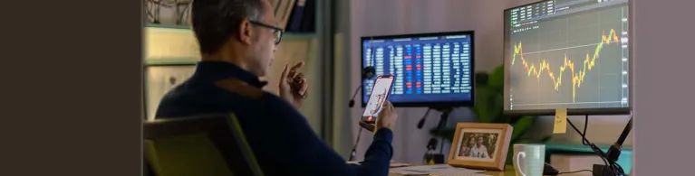 A businessman holds his cellphone to video chat a client overseas while sitting in front of his desk, viewing the foreign exchange stats on the computer screen.