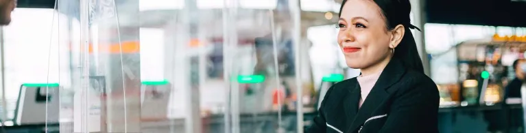 A bank teller stands behind a protective glass shield while handing a checkbook back to a client.