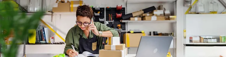 An employee at a small business wearing a green blouse with black overalls smiles as she speaks with a customer on the phone over her office desk.