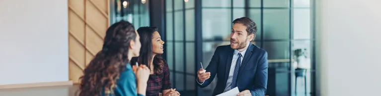 Three professionals in a modern office discuss advanced banking strategies, with documents, a notebook, and a laptop on the table during a collaborative meeting.