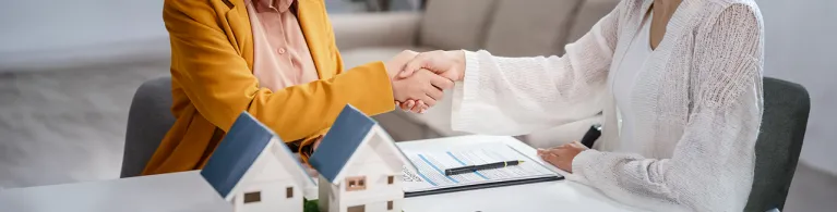 Two individuals sit at a table and shake hands over a mortgage refinancing agreement, with miniature house models, a clipboard of loan documents, and a pen on the table, symbolizing a completed home refinance deal.
