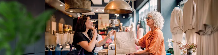 Client and retail worker exchange a shopping bag across a boutique clothing store counter, with racks of apparel and warm lighting in the background.