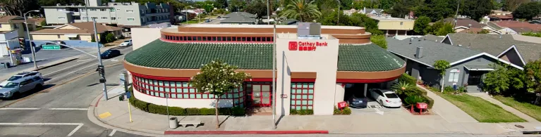 Exterior view of Cathay Bank’s Alhambra Branch, featuring a cream-colored building with a green tile roof, red-framed windows, and prominent Cathay Bank signage. 