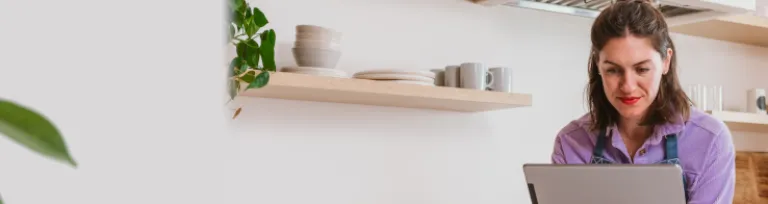 A business woman wearing an apron leans over the counter inside a kitchen looking at her bank account transfers on a digital tablet. 