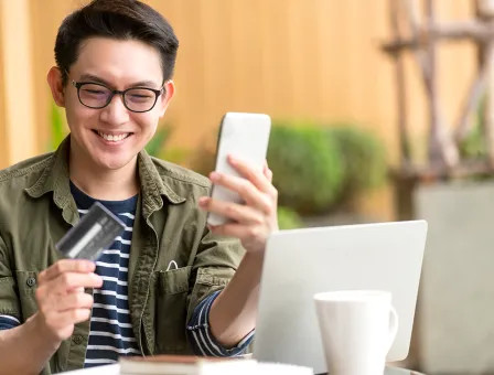  A man is sitting at a table outdoors with a laptop, holding a smartphone in one hand and a credit card in the other, suggesting an online payment or mobile banking activity.