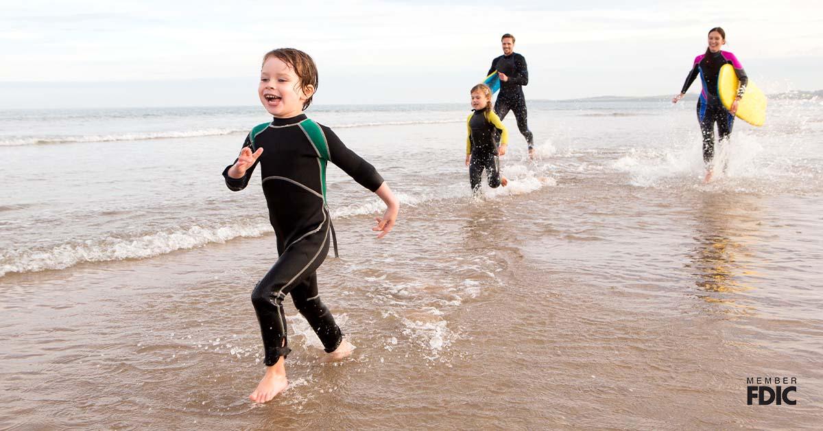 Family of four are running along the shore with body boards. They are all laughing.