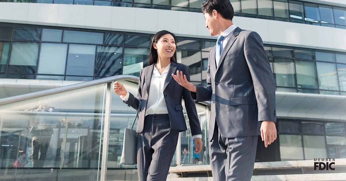 Female professional smiles at her male colleague while walking down the stairs wearing a grey suit, trying to network outside of the office. 