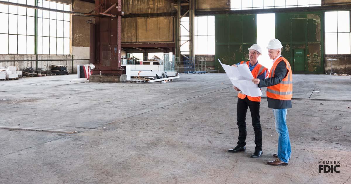 Two men in orange vests and white helmets stand inside a large warehouse holding up construction papers for their next business venture