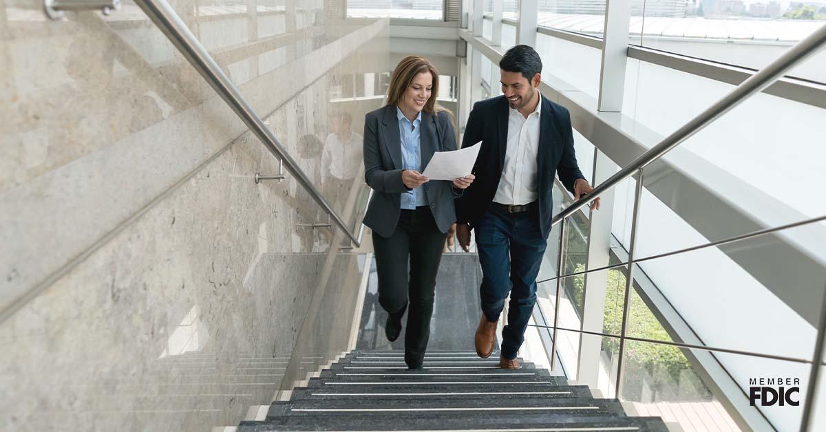 A businesswoman and her male colleague walk up the stairs at an open space office while looking at a business LOC document.